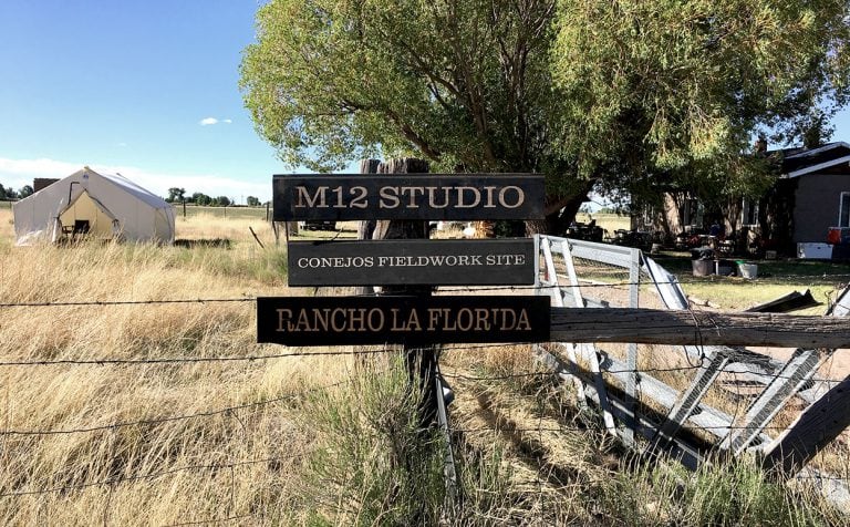 Sign reading: M12 STUDIO / CONEJOS FIELDWORK SITE / RANCHO LA FLORIDA” in front of Adobe Casita and camping tent